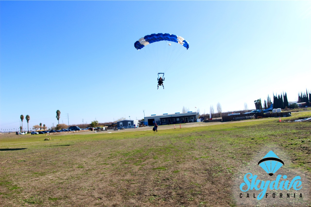 A tandem skydiver and instructor glide smoothly under parachute, preparing for a safe landing in the open field at Skydive California.